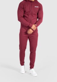 Chándal deportivo burdeos hecho de una tela suave, que incluye una chaqueta con cremallera y pantalones ajustados. Destacado con un logo blanco en el pecho.