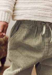 Pantalon en velours côtelé vert clair avec texture côtelée, doté de poches et d'une ceinture à cordon de serrage ajustable, associé à un pull en tricot crème.