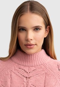 Heled roosa kudumispullover, millel on kõrge ribaga krae ning tekstuurilised mustrid ja avatud töö aktsendid õlgadel.