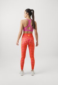 Reggiseno sportivo rosa con design a schiena scoperta, abbinato a leggings elasticizzati con un audace motivo foglia in rosa e arancione, e scarpe da ginnastica bianche.