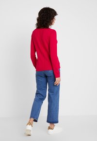 Langärmliges rotes Oberteil mit rundem Ausschnitt, kombiniert mit hellblauen, hoch taillierten Jeans und weißen Sneakers mit einem Akzent im Leopardenmuster.