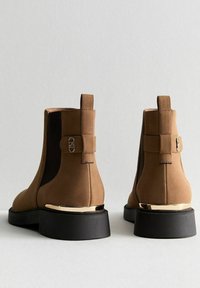 Bottines en daim marron avec semelles en caoutchouc noir, comportant des panneaux latéraux élastiques, des accents dorés sur le talon et une sangle décorative à l'arrière.
