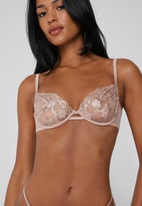 Jasnoróżowy koronkowy biustonosz z kwiecistym haftem, wsparciem z fiszbinami i regulowanymi ramiączkami. Oferuje przezroczyste miseczki oraz dekoracyjny detal na środku.
