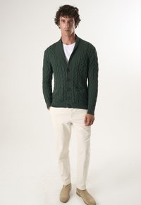 Mørkegrøn kabelstrikket cardigan med sjalkrave, fem knapper og to forlommer, parret med lyse beige bukser og brune støvler.