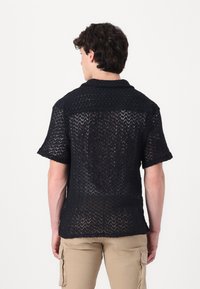 Camisa negra de manga corta hecha de un tejido texturizado de punto abierto con un patrón de chevrón. Presenta un cuello puntiagudo y un corte relajado.