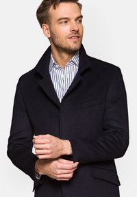 Granatowy wełniany płaszcz z wysokim kołnierzem, pojedynczą kieszenią na piersi oraz guzikami przy mankietach, nałożony na biało-niebieską w paski koszulę.