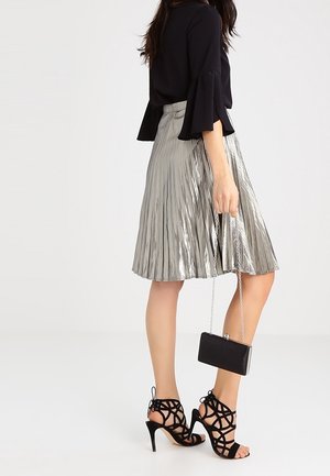 Mujer con falda plisada metálica plateada, blusa negra con mangas acampanadas, zapatos de tacón alto negros con cordones, sosteniendo un pequeño bolso negro con cadena.