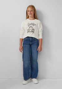 Biała koszula z długim rękawem z grafiką misia pluszowego i napisem "Cuddly Buddy", połączona z szerokimi niebieskimi dżinsami i białymi sneakersami.