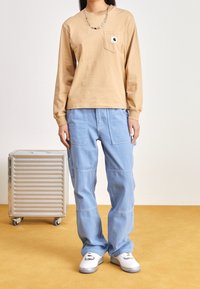 Beige långärmad skjorta med ficka och logotyp, kombinerad med blå cargobyxor. Vita sneakers med svarta accenter, texturerad bakgrund.