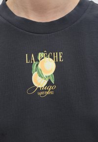 Μαύρο πουκάμισο με κίτρινο κείμενο "LA PÊCHE Hugo SAINT-TROPEZ" και δύο εικονογραφημένα ροδάκινα με πράσινα φύλλα κάτω από το κείμενο.