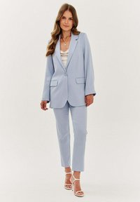 Světle modrý oblek na míru s blejzrem na jedno knoflíkové zapínání a rovnými kalhotami. Hladká látka, dvě přední kapsy, doplněné bílými sandály na pásek.