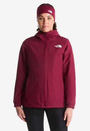 Kvinde iført en mørkerød hættejakke og matchende pandebånd med The North Face-logoer, stående mod en hvid baggrund.