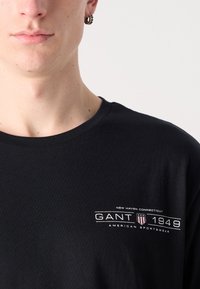 Black cotton T-shirt with a crew neck, featuring a white logo print that includes "GANT 1949" and "New Haven Connecticut."