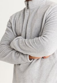 Grå fleecebluse med høj hals, fuld lynlås og tekstureret overflade. Har lange, tætsiddende ærmer og en afslappet pasform.