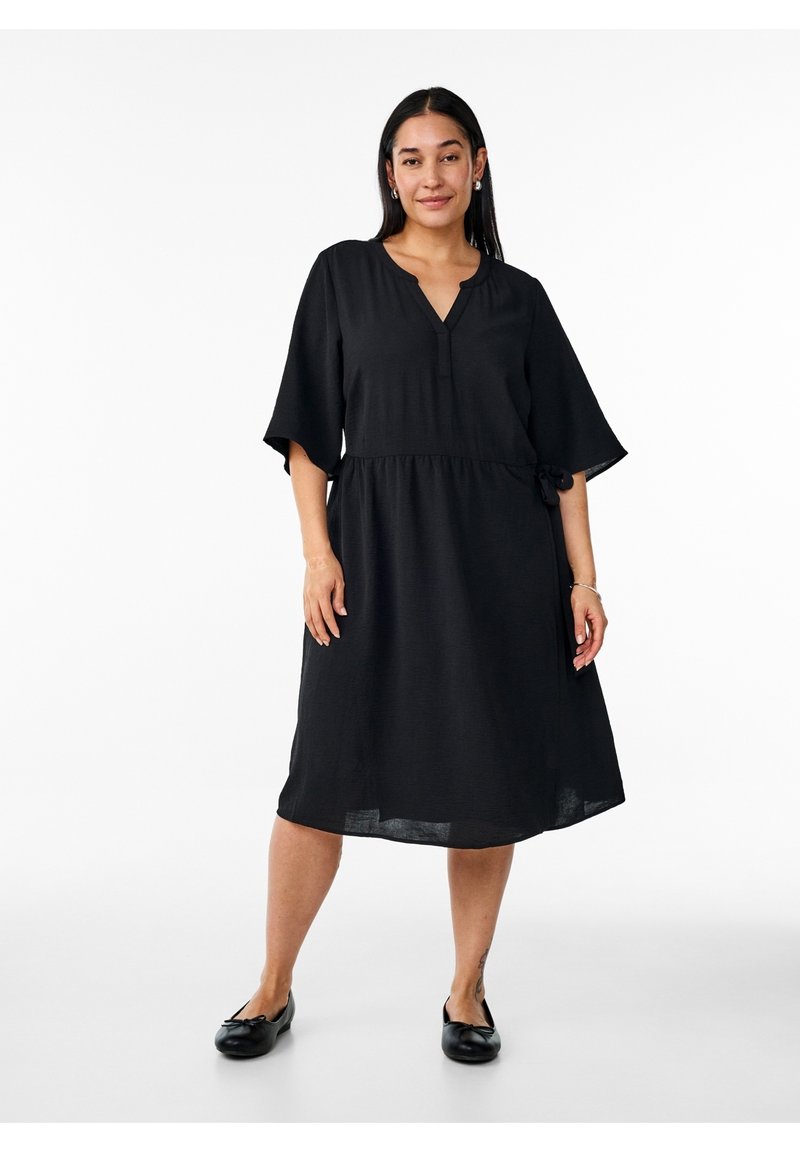 Femme aux longs cheveux foncés portant une robe midi noire ample avec des manches au coude et des chaussures plates noires, debout sur un fond blanc.