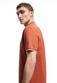 Kopparfärgad polo med en strukturerad tyg, som har en vit kant runt kragen och ärmarna. Modellen poseras i profil.