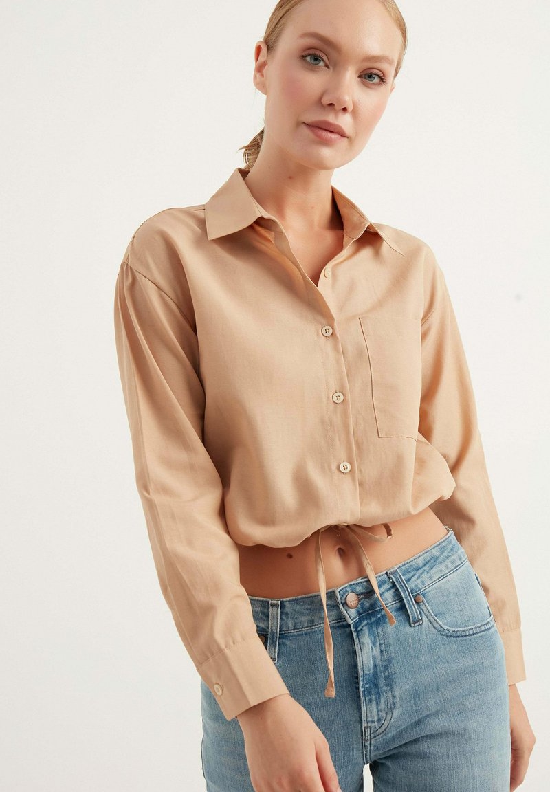 Camicia cropped button-up color beige chiaro con maniche lunghe, una tasca sul petto e un dettaglio a nodo sull'orlo, abbinata a jeans blu a vita alta.