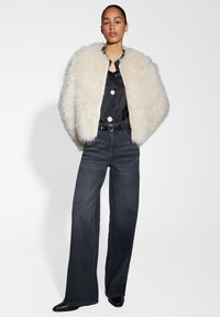 Giacca in pelliccia sintetica crema, camicia nera in satin con bottoni, jeans a gamba larga scuri e scarpe nere a punta contro uno sfondo semplice.