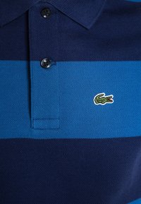 Navy- und hellblau gestreiftes Poloshirt aus strukturiertem Material, mit einer Zwei-Knopf-Leiste und einem kleinen grünen Krokodil-Logo auf der Brust.