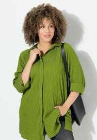 Camicia verde, texturizzata, con bottoni e colletto, con le maniche arrotolate. Indossata con jeans scuri e una borsa a spalla nera. Design semplice.
