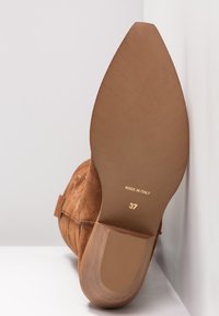 Brun ruskindsankelstøvle med afrundet hæl, der har en glat sål og gyldne prægede mærker: "Made in Italy" og størrelse "37".