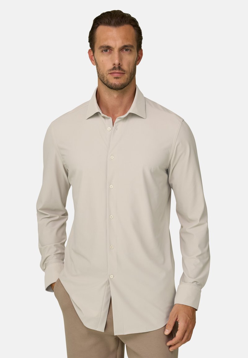Homme aux cheveux courts châtain, portant une chemise beige à manches longues boutonnée et un pantalon marron clair, debout avec une main dans la poche.