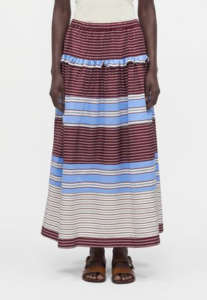 Femme portant une longue jupe rayée avec des bandes horizontales bordeaux, bleu clair et blanches, et des sandales marron, debout devant un fond uni.