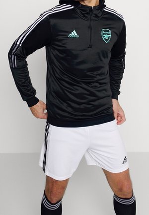 Männlich, trägt schwarzes Adidas Arsenal Trainingsoberteil mit weißen Streifen und weiße Adidas Shorts mit schwarzen Streifen, posiert mit den Händen auf den Hüften.