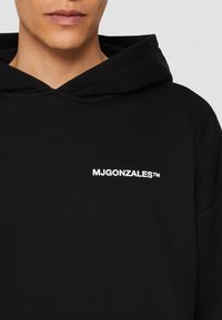 Un sweat à capuche noir avec un cordon de serrage et un petit logo blanc "MJGONZALES™" sur la poitrine gauche. Texture de tissu doux et côtelé.