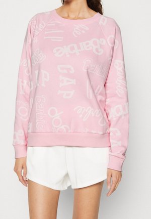 Kobieta ubrana w różową bluzę z białymi, rozrzuconymi logo tekstowymi oraz białe szorty, stojąca na tle jednolitego, jasnego tła.