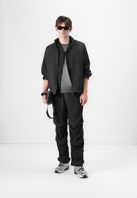 Veste zippée noire sur un t-shirt gris, associée à un pantalon noir et des baskets blanches. Les accessoires comprennent des lunettes de soleil noires et un petit sac.