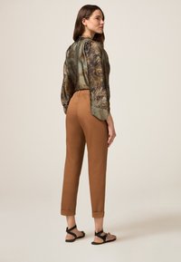 Groene blouse met een patroon en losse mouwen, gecombineerd met bruine taps toelopende broek en zwarte sandalen, die een ontspannen pasvorm en zachte texturen tonen.