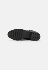 Suela de zapato negra de material de goma grueso, con un patrón en zigzag para tracción y un logo en relieve en el centro.