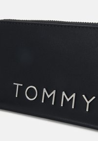 Černá syntetická taštička s texturovaným povrchem a stříbrným logem "TOMMY". Obsahuje zip a kontrastní švy.