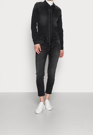 Person iført sort denimjakke og jeans med hvid rullekrave og hvide sneakers, står på et almindeligt gulv mod en hvid væg.