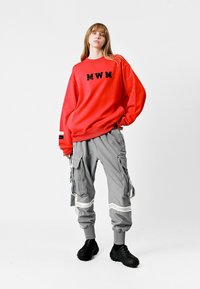 Sudadera roja con "MWM" en lentejuelas negras, combinada con pantalones cargo grises que tienen múltiples bolsillos y detalles en rayas blancas. Zapatos negros.