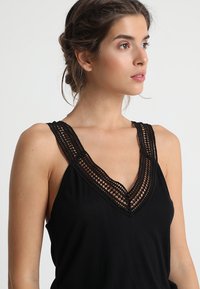 Black tank top featuring a deep V-neck with intricate lace detailing along the neckline. Smooth fabric with a relaxed fit and sleeveless design.