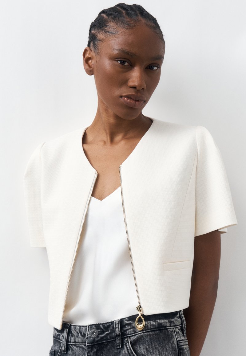 Γυναίκα με πλεξούδες φοράει λευκό, κοντομάνικο, κοντό μπουφάν με χρυστό φερμουάρ πάνω από άσπρο τοπ και μαύρα ξεβαμμένα τζιν.