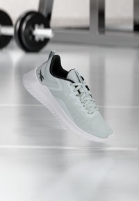 Helle graue Sportschuh mit Obermaterial aus Mesh, texturierten Details, flachen Schnürsenkeln und einer weißen Gummisohle, vor einem verschwommenen Fitnessstudio-Hintergrund abgebildet.