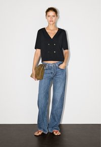 Schwarze, kurzärmelige, bauchfreie Bluse mit doppelten goldenen Knöpfen, kombiniert mit blauen, weit geschnittenen Jeans und einer braunen Clutch; flache rote Sandalen.
