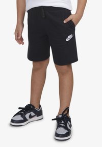 Fekete pamut rövidnadrág, rugalmas derékpánttal, oldalzsebbel és fehér Nike logóval; fekete és szürke sportcipőkkel párosítva.