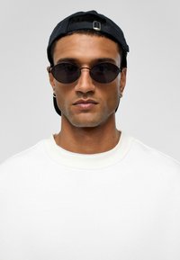 Zwarte ovale zonnebril met dunne zwarte frames. Het model draagt een witte sweatshirt en een zwarte pet, met een gebogen rand en een metalen verstelmechanisme.