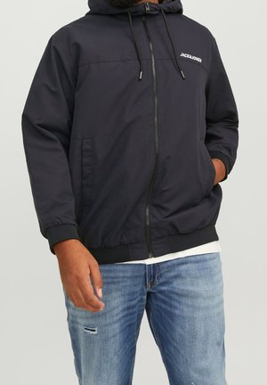 Mand iført sort lynlåshættejakke med "JACK & JONES" logo på brystet, kombineret med lyseblå slidte jeans.