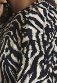 Schwarz-beige gemusterte Bluse mit gerafften, strukturierten Ärmeln und einem auffälligen Tierdruckdesign. Weicher Stoff mit glatter Oberfläche.