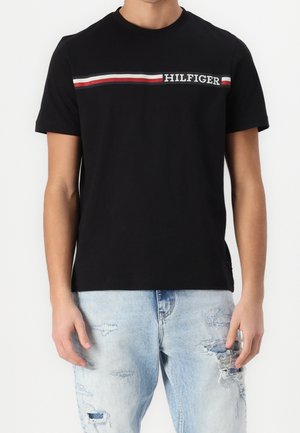 Camisa negra de manga corta con franjas horizontales en rojo, blanco y negro, y el texto "HILFIGER", combinada con vaqueros claros desgastados.