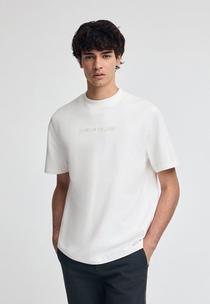 Camiseta de algodón blanca con mangas cortas, cuello redondo y logo en relieve "POLO CLUB". Corte holgado con una textura suave y sin patrones visibles.