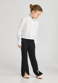 Camicia bianca con bottoni e maniche lunghe, abbinata a pantaloni neri con fessure laterali. Il modello indossa ballerine rosa chiaro.