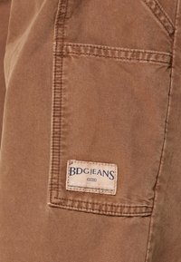 Jeans de algodón marrón con una tela texturizada, costuras visibles y una etiqueta de tela que dice "BDGJEANS" en el bolsillo trasero.
