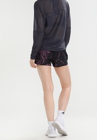 Musta urheilushortsit, joissa on vaaleanpunainen abstrakti kuvio, yhdistetty tumman pitkähihainen paita. Malli pukeutuneena valkoisiin sukkiin ja lenkkareihin.