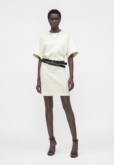 Γυναίκα στέκεται, φοράει ένα κρεμ μίνι φόρεμα με κοντά μανίκια, μαύρη διπλή ζώνη, ασημένιο αλυσίδα κολιέ και μαύρα σανδάλια με ψηλό τακούνι.